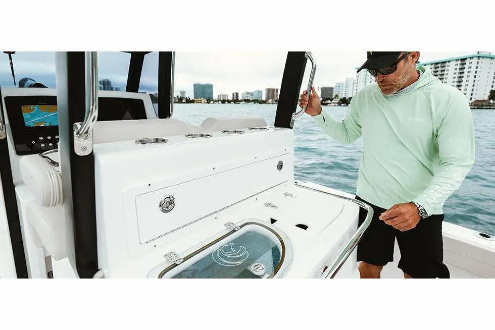 A man in a light green shirt operates a boat control panel with city buildings and water in the background.